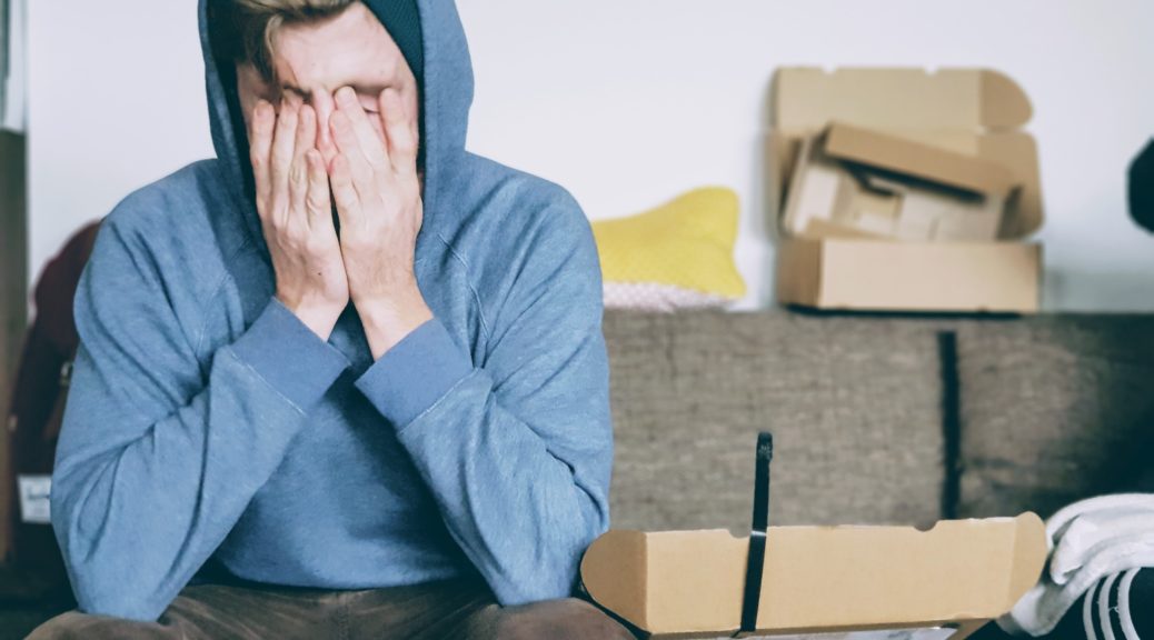 Man wearing a hoodie covering face with both hands while sitting on couch