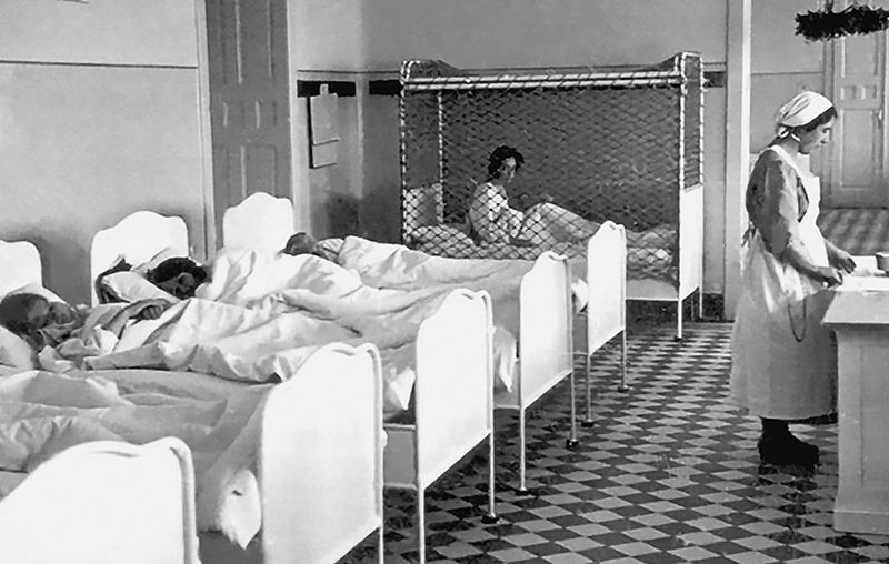 A black-and-white photograph depicting children in a row of beds, one of which is enclosed by a mesh cage. A woman in a nurse's uniform stands facing away from the beds.
