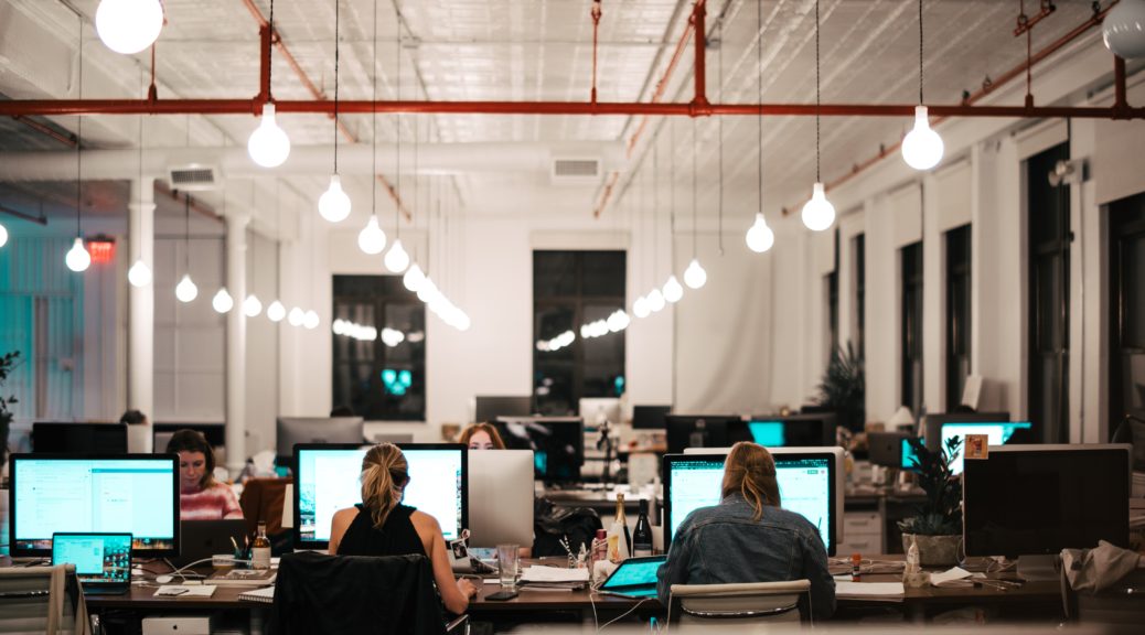 A busy office with people sitting in front of computer monitors. Work areas are not separated by cubicles. Illumination is provided by exposed light bulbs hanging overhead.