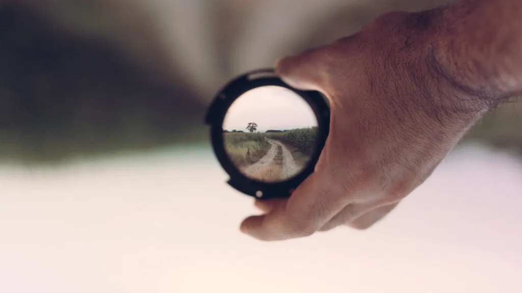 A hand, surrounded by blurry scenery, holding a lens through which an upside-down and sharper image of the area ahead is visible.